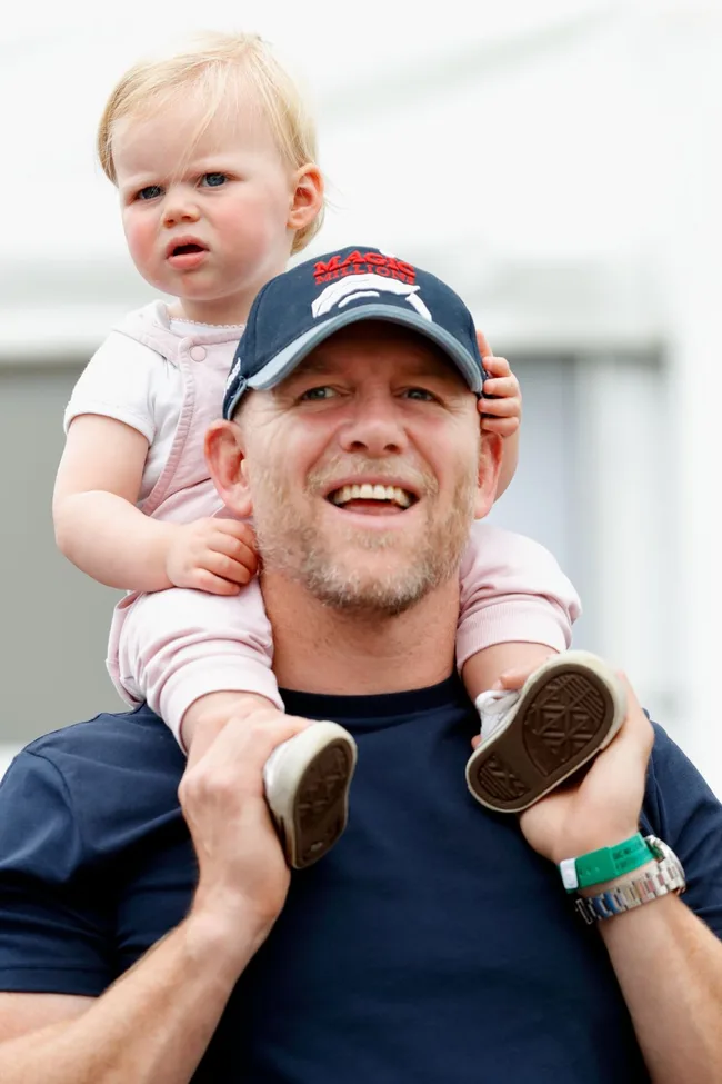 Man smiling with a cap, holding a toddler on his shoulders outside. Toddler looks curious and is wearing pink.