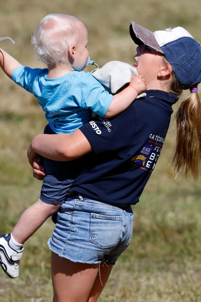 A girl in a cap holds a toddler with a pacifier and soft toy in a field.