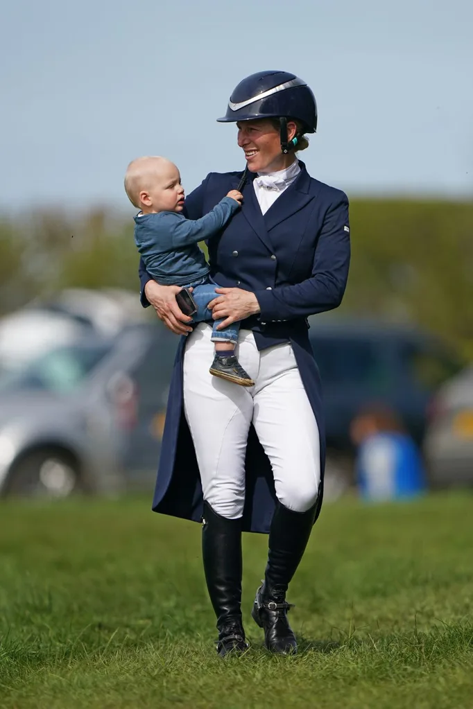 Woman in equestrian attire holds a baby, smiling outdoors on a sunny day.