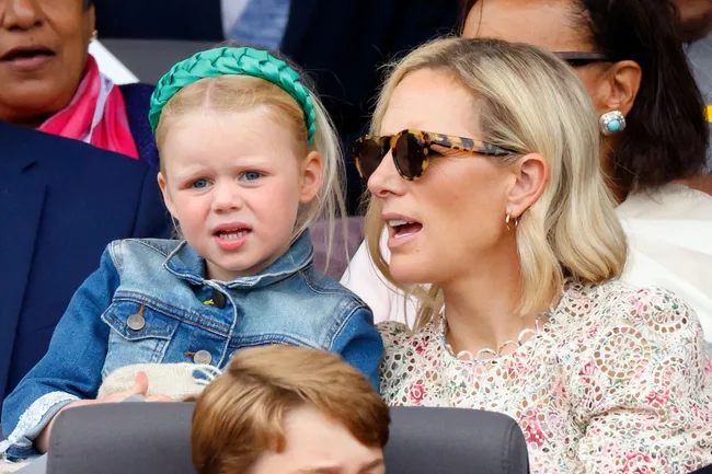 Woman in sunglasses and floral top with a child in a denim jacket and green headband at an outdoor event.