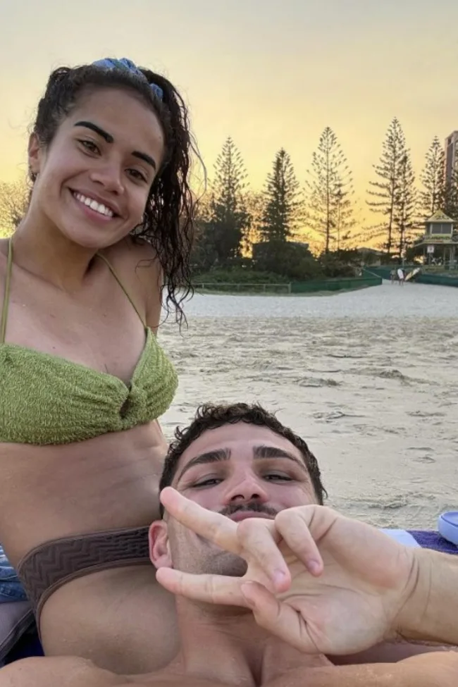 Two people pose on a beach at sunset; the woman smiles, and the man shows a peace sign.