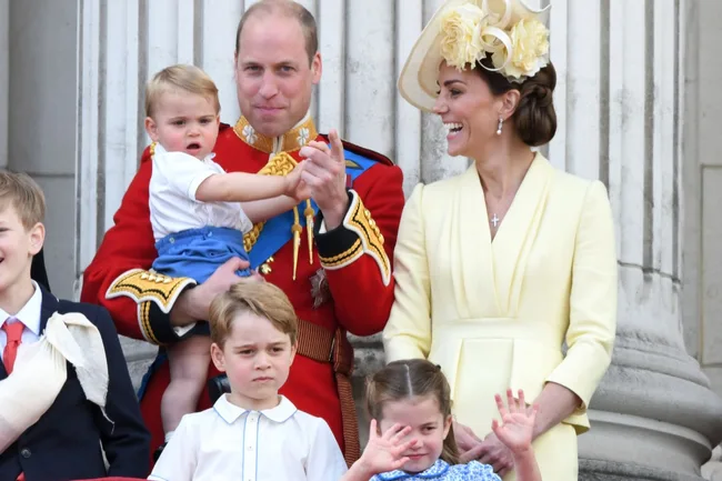 Royal family on balcony; man in uniform, woman in hat, and three children, one waving, smiling and interacting.