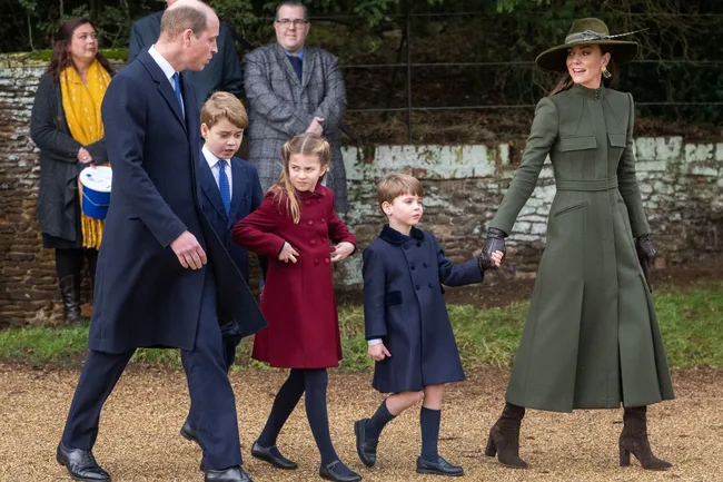 Royal family walking, with woman in green coat holding hands with young boy in blue coat, and man leading other children.