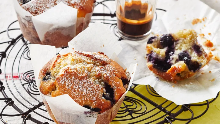 Freshly baked blueberry muffins dusted with powdered sugar on a wire rack, with coffee in the background.