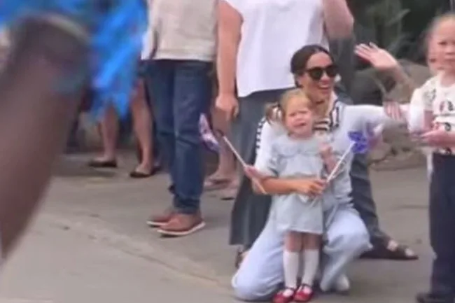 A woman in sunglasses kneels, smiling with a young girl holding a pinwheel, surrounded by a crowd.