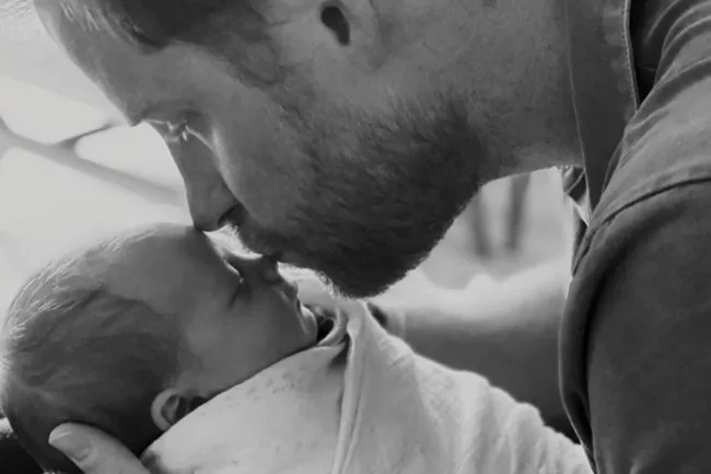 A man gently kisses the forehead of a swaddled baby in a tender black-and-white photograph.