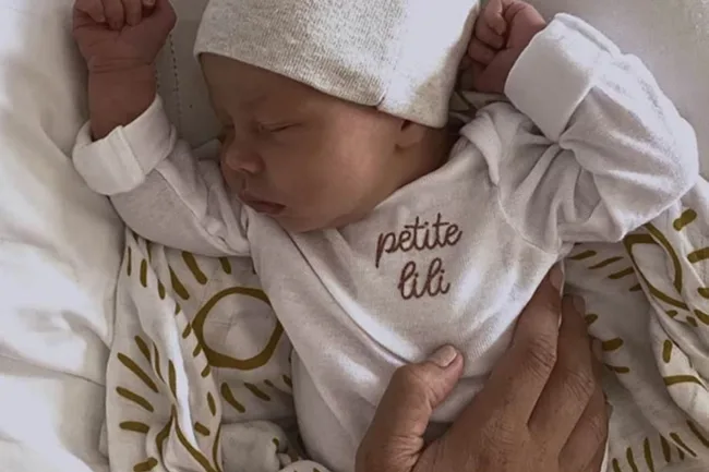 Sleeping baby in a white onesie with "petite lili" text, wearing a matching hat, held by an adult's hand.