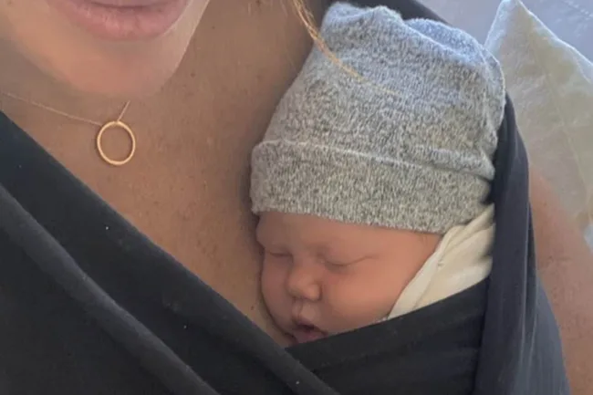 A baby wearing a gray beanie sleeps snugly against a person's chest wrapped in a black fabric sling.