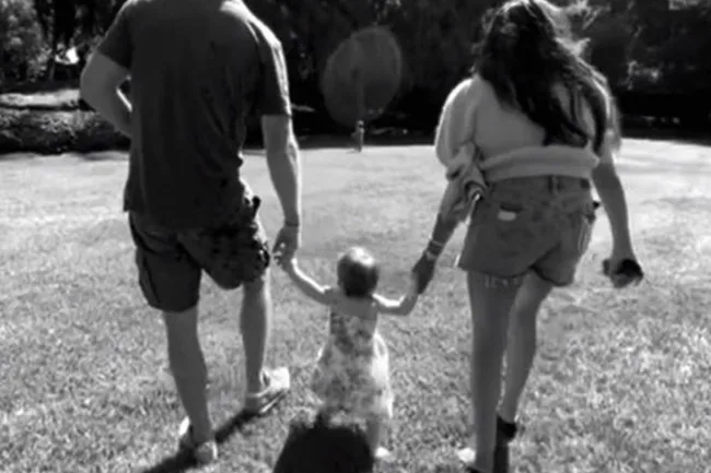 Black and white photo of two adults walking hand-in-hand with a toddler on grass.