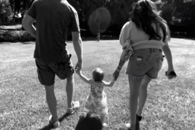 Black and white photo of two adults walking hand-in-hand with a toddler on grass.