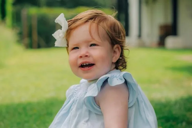 A young child with red hair and a white bow in a blue dress, smiling outdoors on a bright, grassy day.