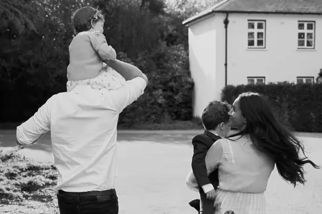 A family walking away; a man carries a child on his shoulders, a woman holds another child, with a house in the background.