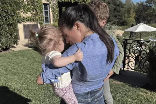 A woman in a denim shirt holds a young child outdoors beside a railing and garden.