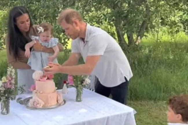 A child being held by a woman as a man decorates a two-tier cake outdoors, with flowers on the table.