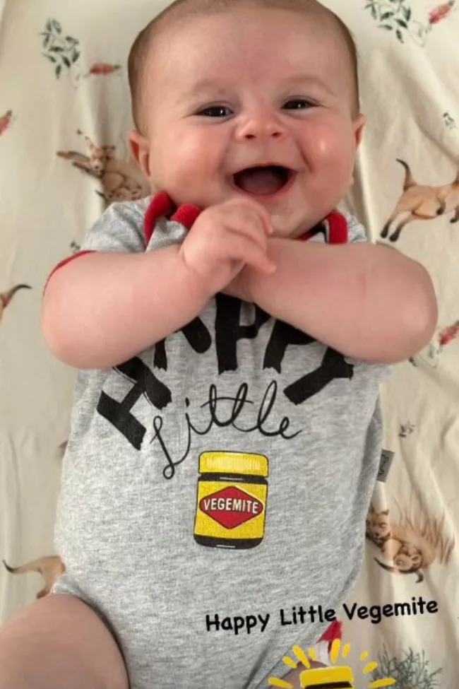 Smiling baby wearing a onesie with "Happy Little Vegemite" text and Vegemite jar graphic.