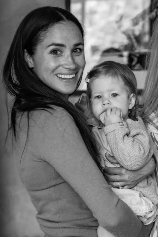 A woman smiling while holding a young child who is biting their hand, both in a cozy indoor setting, black and white photo.