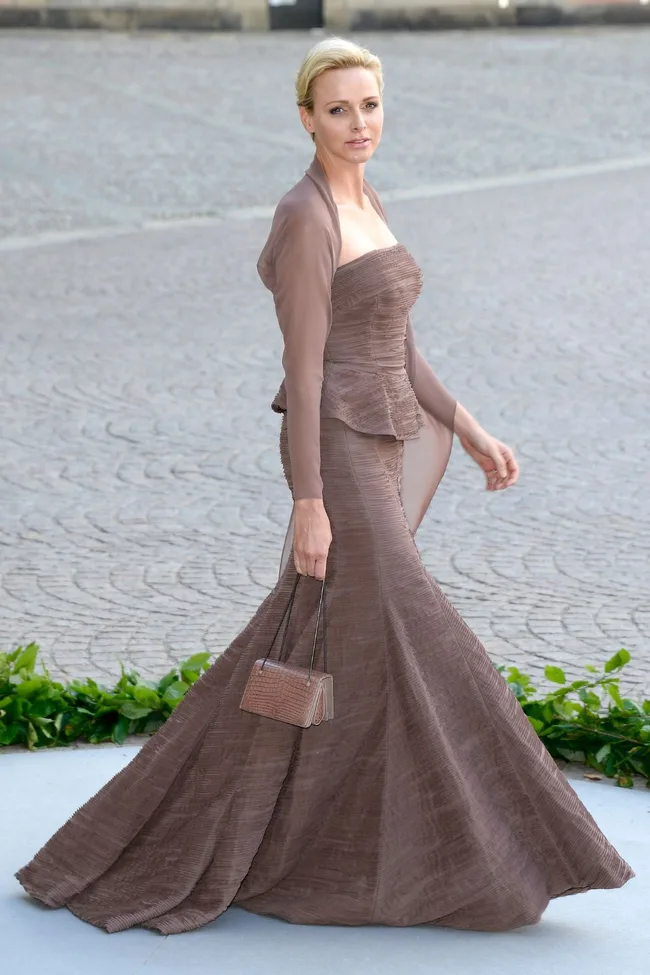 Woman in elegant brown gown walking on a cobblestone path, carrying a small handbag.