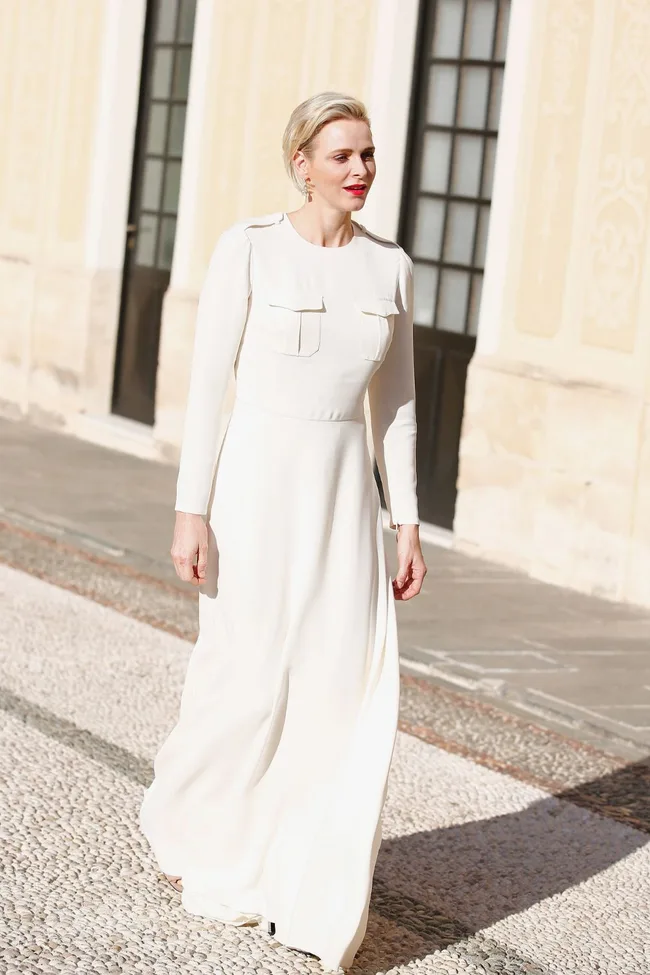 A woman in a long white dress walks outside a building with beige walls and large windows.
