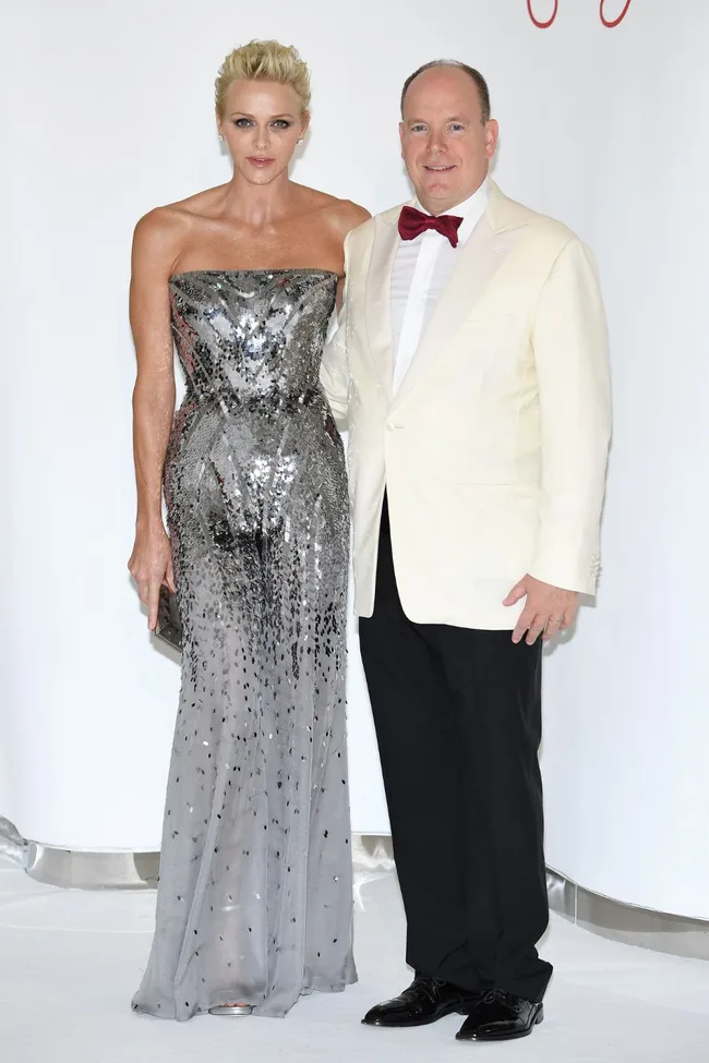 A couple posing elegantly; woman in silver sequin gown and man in white tuxedo with a bow tie on a white backdrop.