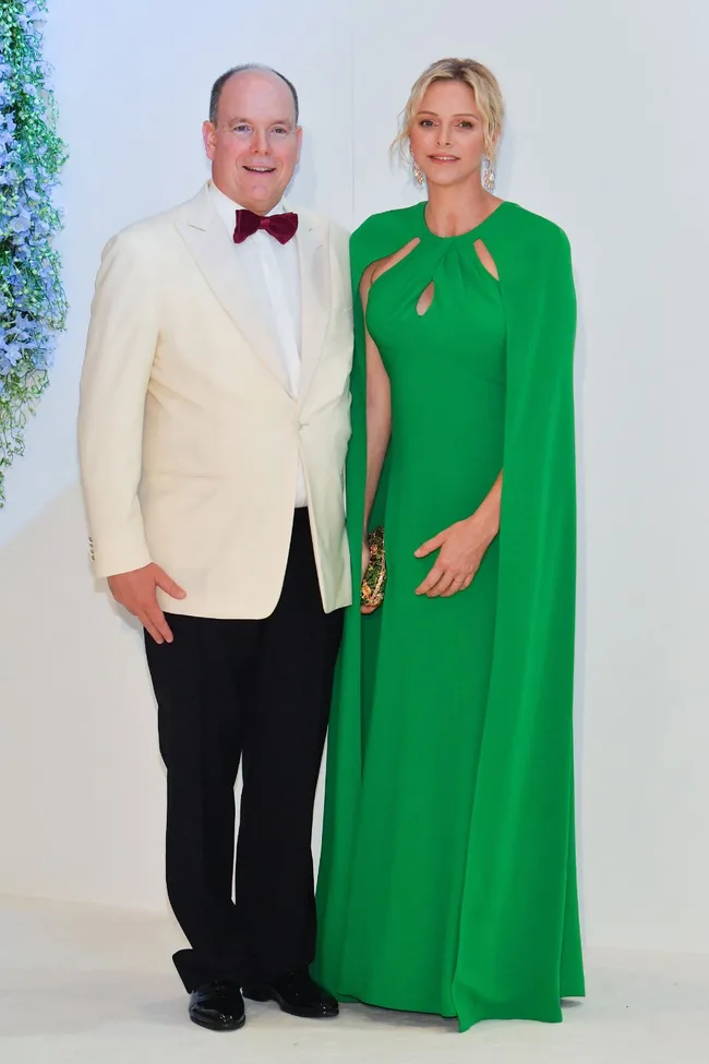 A man in a white tuxedo and woman in a green gown pose together at an event against a light background.