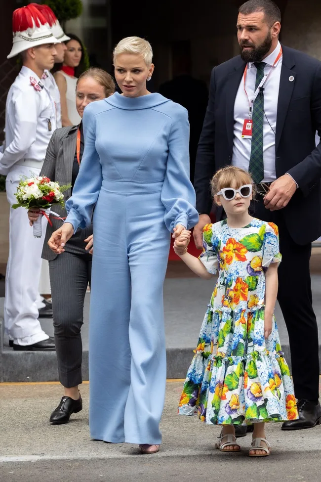 A woman in a blue outfit holds hands with a girl in a floral dress and sunglasses, with a security detail nearby.