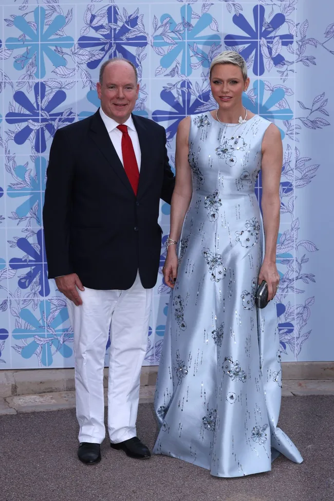 A man in a suit with a red tie and a woman in a silver floral gown pose in front of a blue patterned wall.