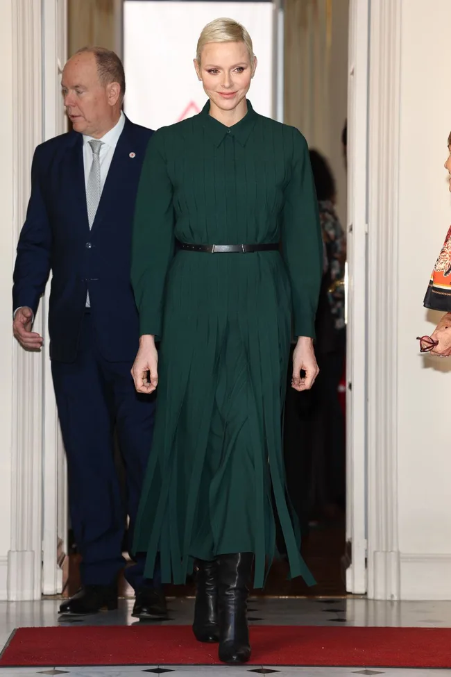 Elegant woman in a green dress and black boots walks confidently, with a man in a suit behind her.