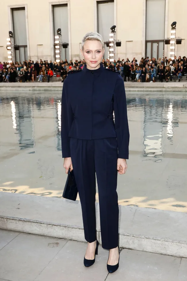 A woman in a navy suit stands by a reflective pool, with an audience in the background.
