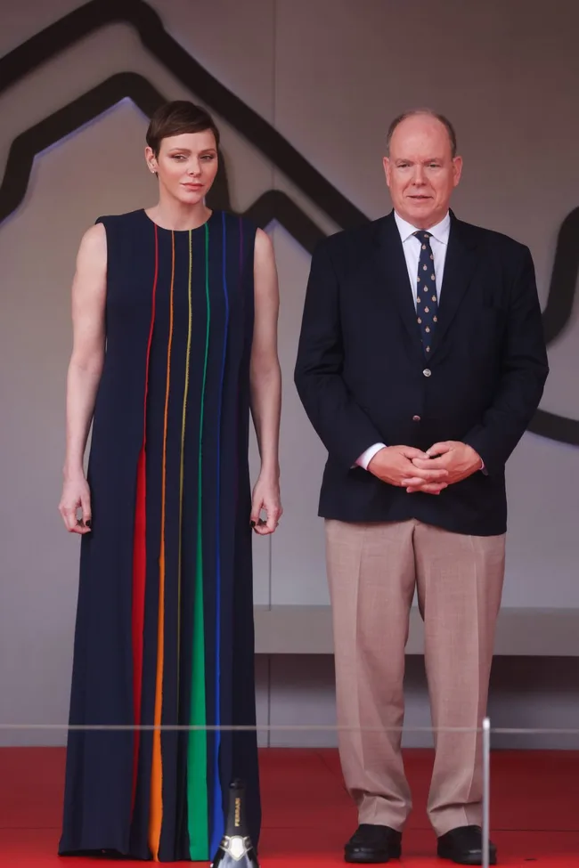 Two people stand on a red carpet; one in a multicolored dress and the other in a suit and tie.