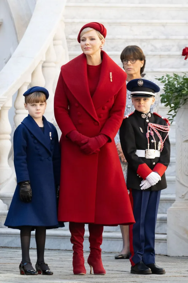 Three individuals pose on stairs; woman in red coat and hat, child in blue coat, and child in uniform.