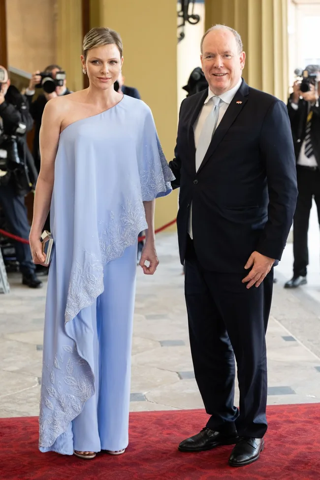 Man and woman pose elegantly on a red carpet, woman in light blue one-shoulder outfit, photographers in background.
