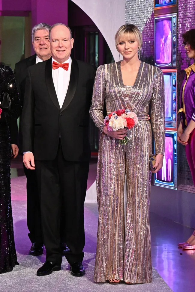 A couple in formal attire, the woman in a metallic jumpsuit holding flowers, standing at a glamorous event.