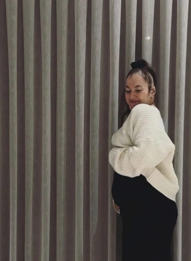 A pregnant woman in a white sweater stands sideways, smiling, with a background of vertical curtains.