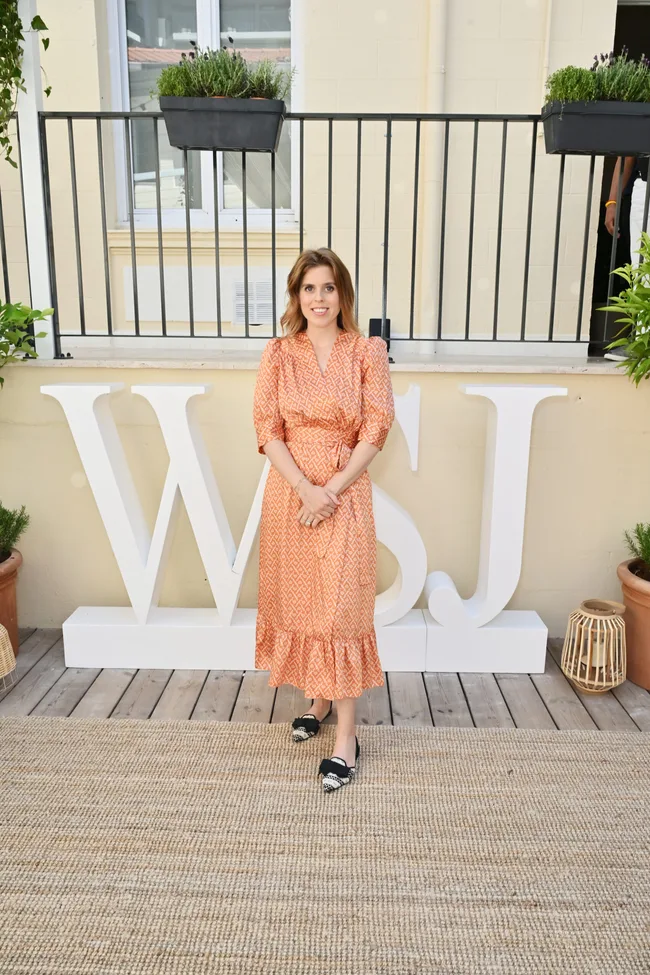 Princess Beatrice in an orange patterned dress at a WSJ event, standing on a deck with potted plants.