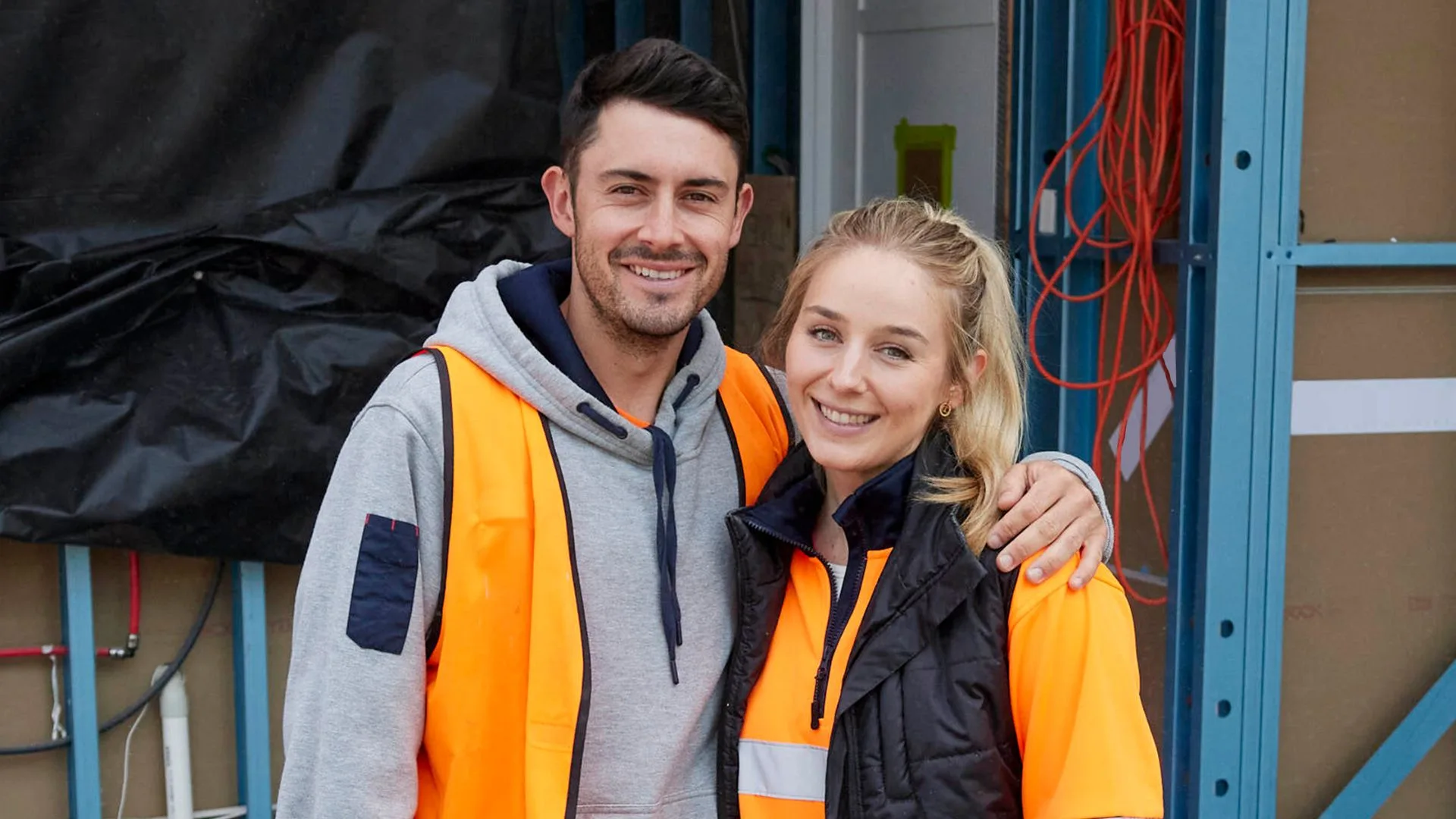 Man and woman in orange safety vests smiling at a construction site.