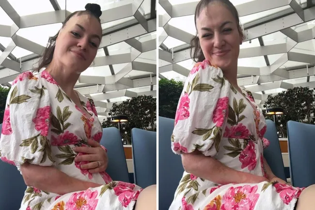 Woman in floral dress sitting, smiling, and holding her belly in a bright, modern indoor setting.