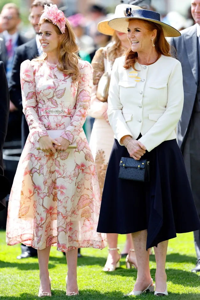 Princess Beatrice in a floral dress and fascinator beside another woman in a white jacket and navy skirt, both smiling outdoors.