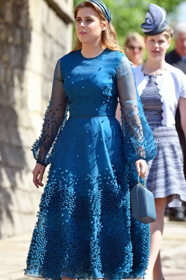 Woman in a blue textured dress with sheer sleeves, holding a matching handbag, walking outdoors.