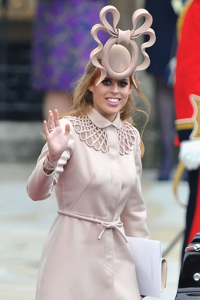Princess Beatrice in a blush coat and dramatic fascinator, smiling and waving at a public event.