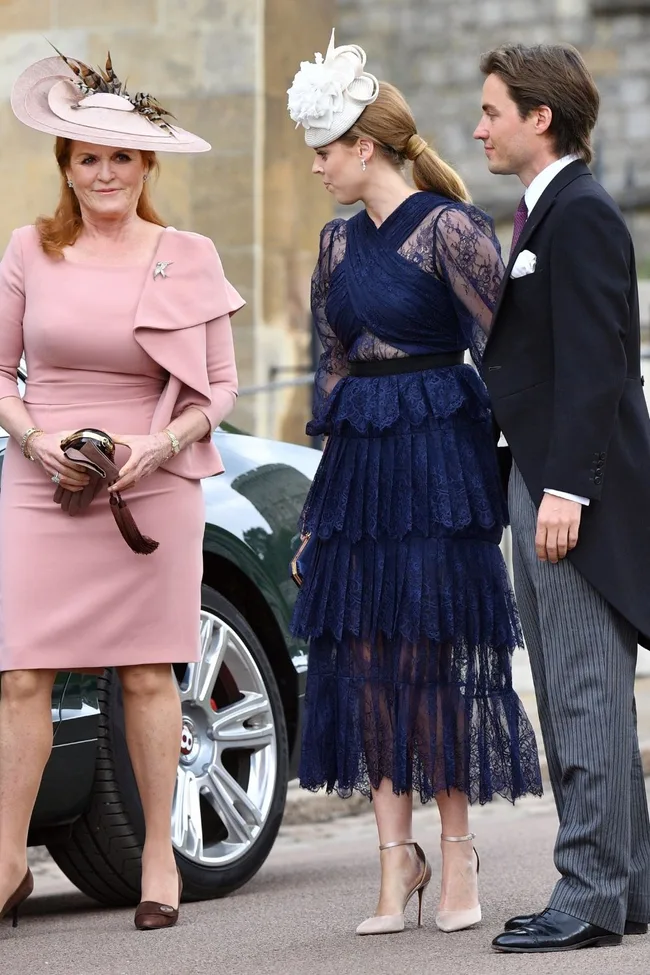Three people dressed in formal attire, one in a pink dress and hat, another in a blue lace dress, outside a classic car.
