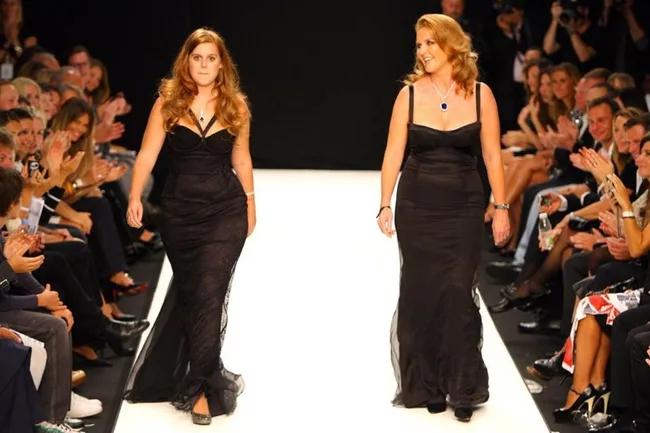 Two women in black gowns walk a runway, audience clapping on both sides.
