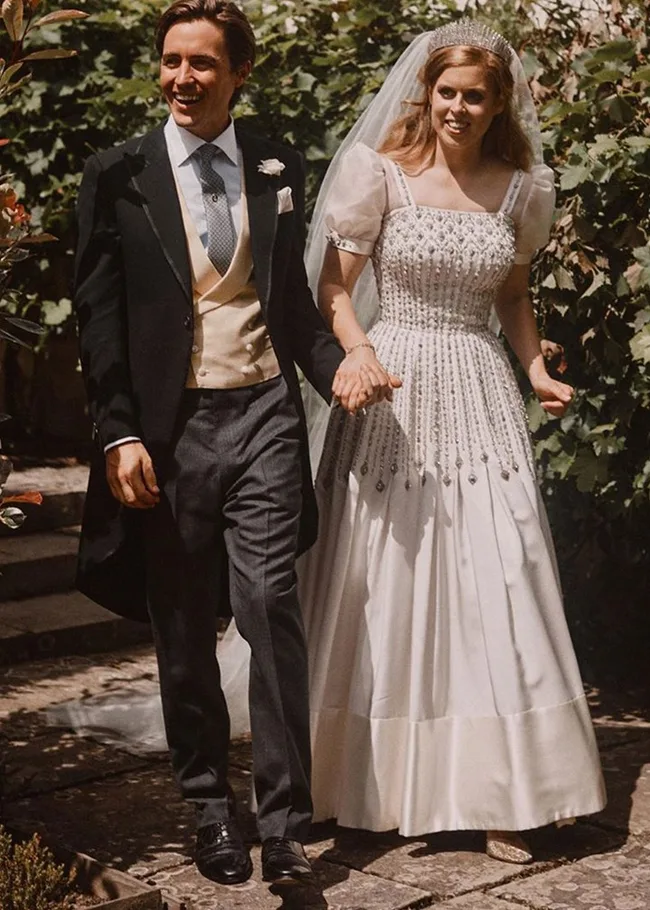 Bride in a vintage wedding dress with puff sleeves and tiara, holding hands with groom in a formal suit, walking outdoors.