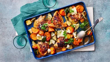Baked chicken thighs with roasted vegetables in a blue tray, garnished with herbs.