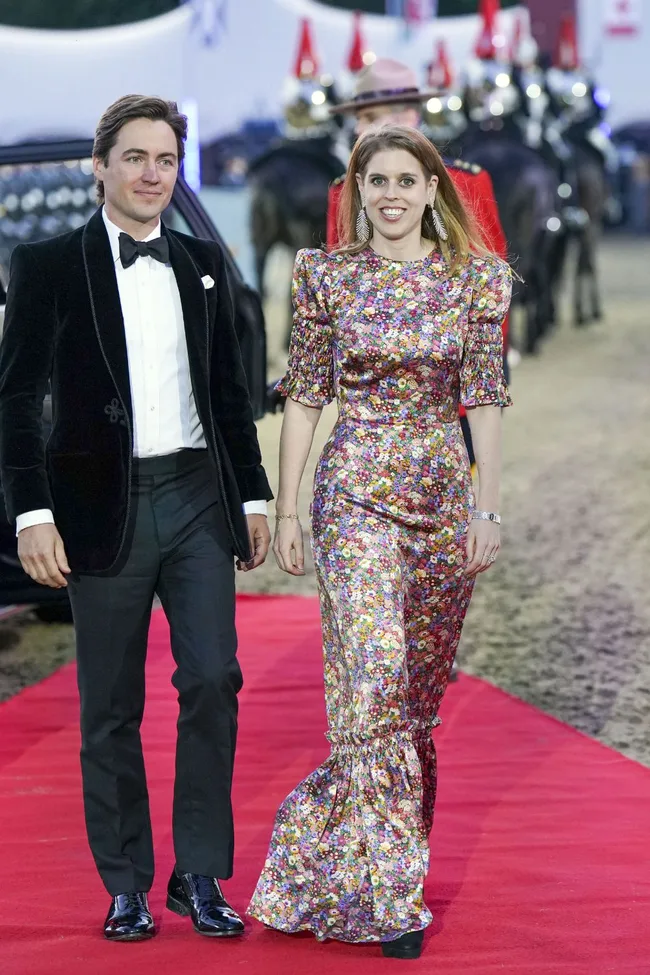 A couple walks on the red carpet, man in a black tuxedo and woman in a floral dress, with an equestrian guard in the background.