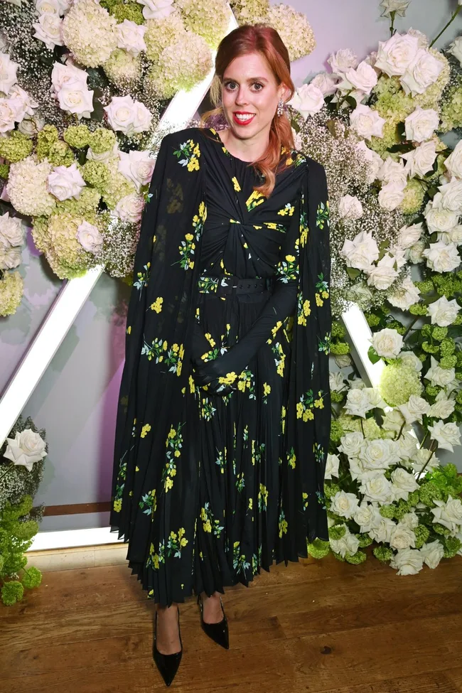 Princess Beatrice in a floral black dress stands in front of a floral backdrop.