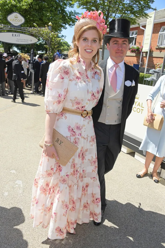 Princess Beatrice at Royal Ascot in a floral dress, matching hat, and holding a monogrammed clutch, posing with a companion.