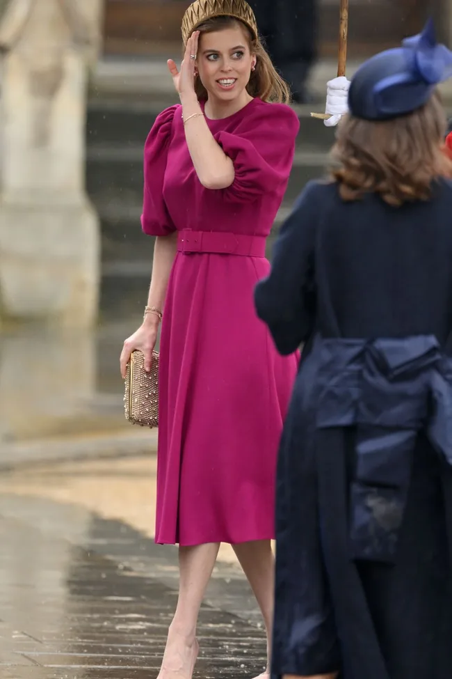 A woman in a magenta dress and gold headband holds a clutch, interacting with another person wearing a navy outfit.