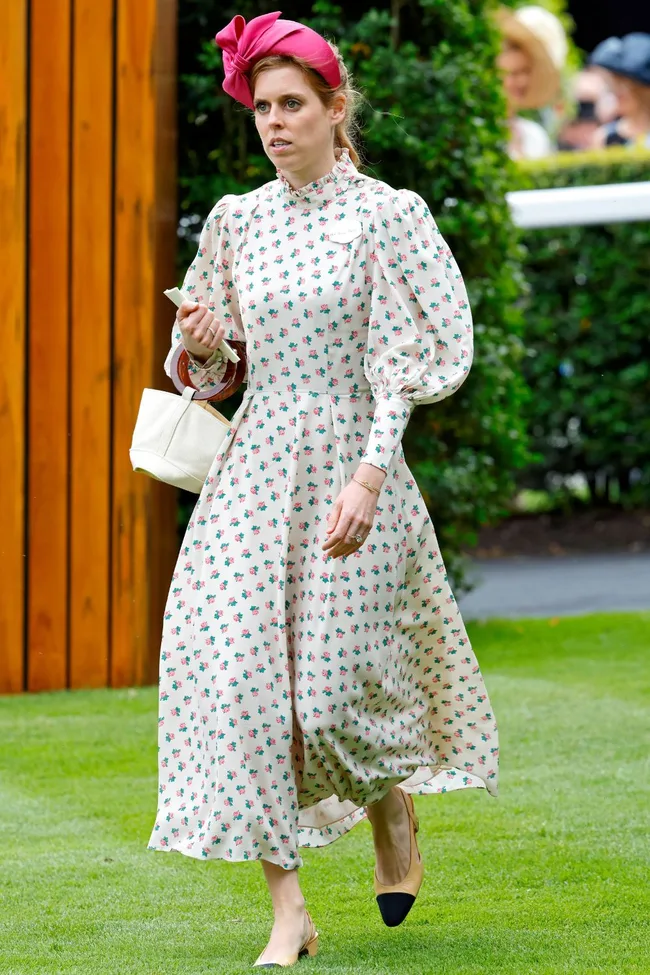 A woman in a floral dress and pink headband walks on grass, holding a white bag.