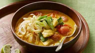 A bowl of vibrant Thai curry soup with noodles, chicken, tomatoes, green onions, and lime slices on the side.