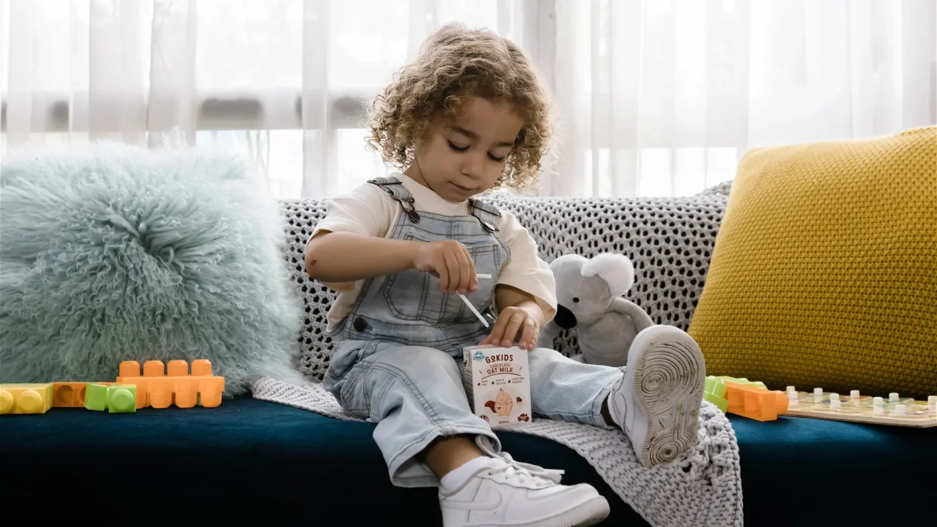 Child in denim overalls drinking oat milk on a couch, surrounded by toys and stuffed animals.